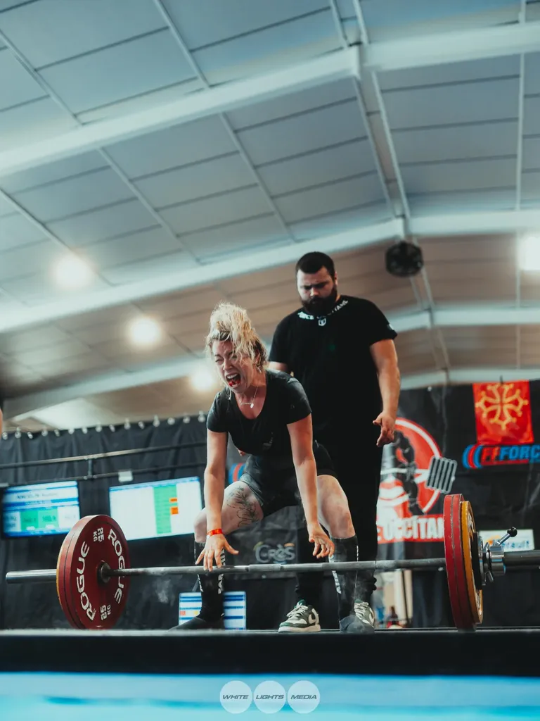 Une athlète crie en s'approchant de la barre de soulevé de terre, son coach juste derrière elle, sur un plateau FFForce. Photo @audrey_boucle / @whitelightsmediafr.